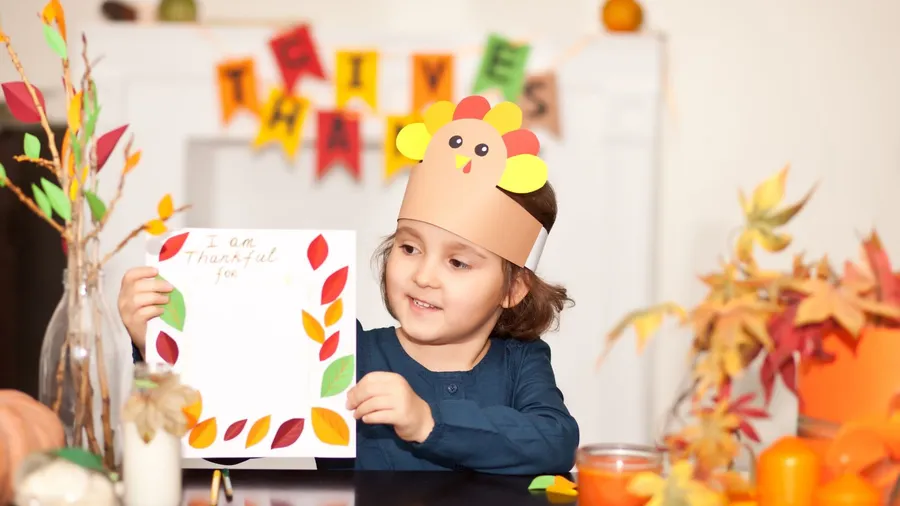 A young girl wearing a paper turkey hat, holding a thankful message card, with fall decorations.