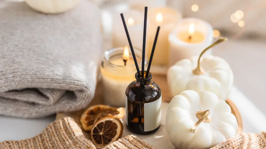 Close up of white pumpkins, candles, dried citrus slices, a reed diffuser, and a folded blanket placed on a white table.