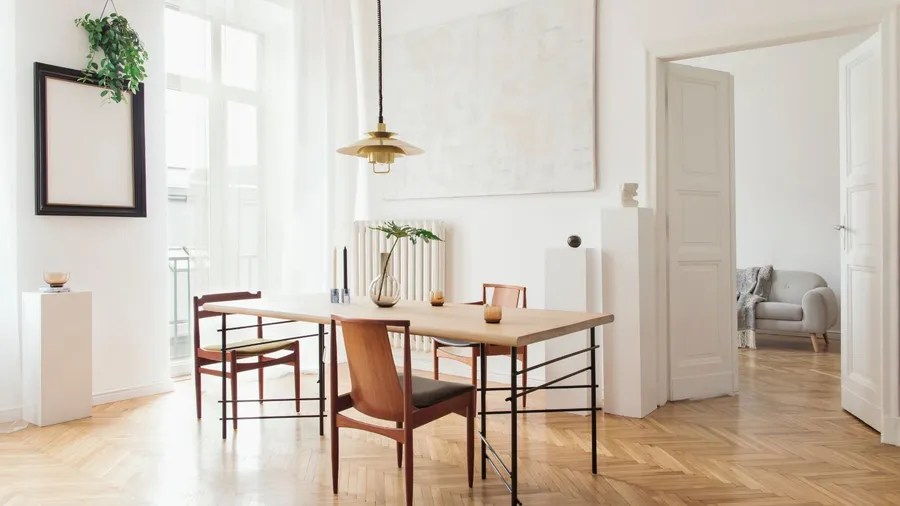 A bright dining room featuring a wooden table surrounded by chairs and plants against a white background.