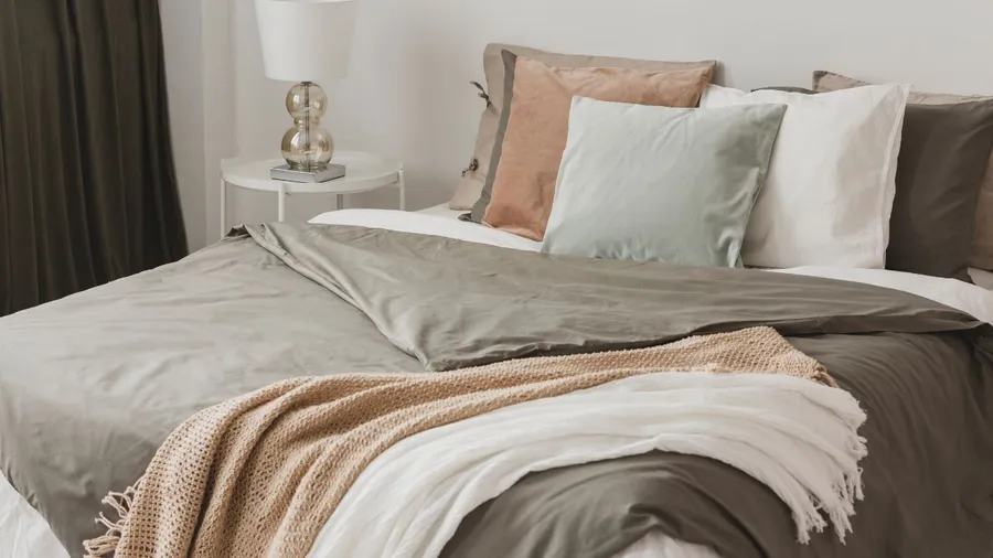 A neatly made bed with gray and white bedding, multiple pillows, and a bedside table with a lamp.