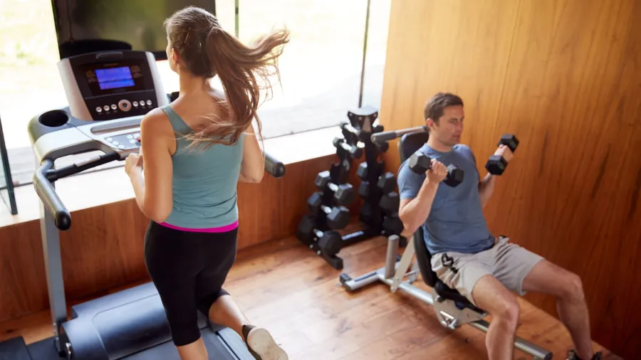 A woman jogs on a treadmill, while a man lifts dumbbells in a gym.