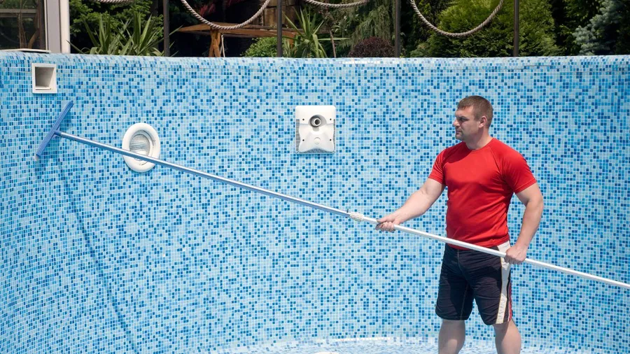 Person cleaning a blue mosaic-tiled pool with a long pole.