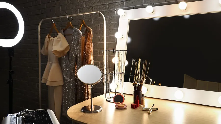A well-lit vanity station with a large mirror, makeup brushes, and other beauty essentials. And a hanging rack with dresses.