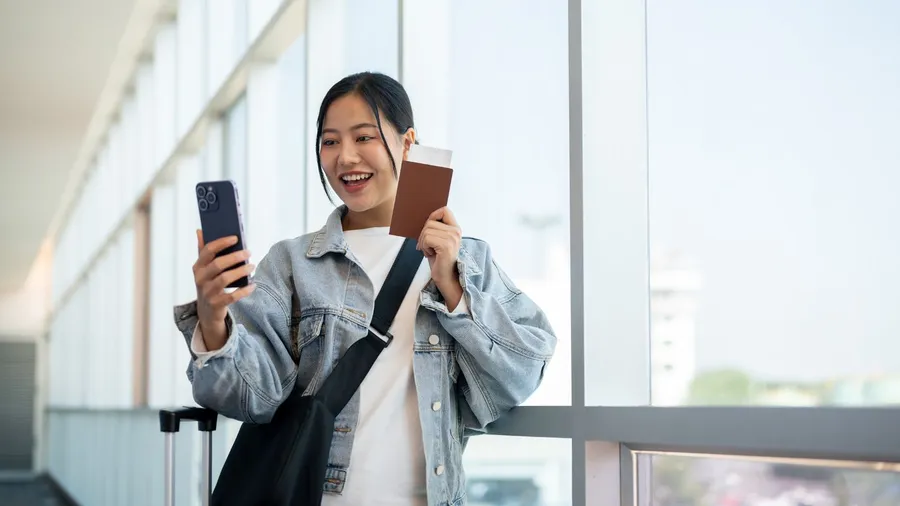 A cheerful, excited Asian female passenger enjoys talking on a video call or recording a video on her smartphone while waiting for her flight boarding time.
