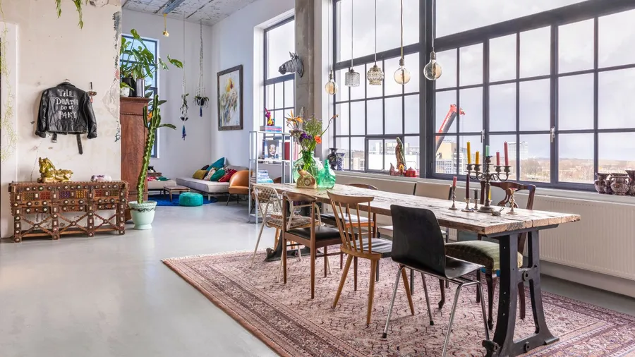 An eclectic dining area showcases a rustic table, diverse decor, and large industrial windows for a vibrant atmosphere.