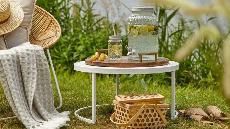 A patio scene with a chair, a table holding a glass jar of water, and a picnic basket on the grass.
