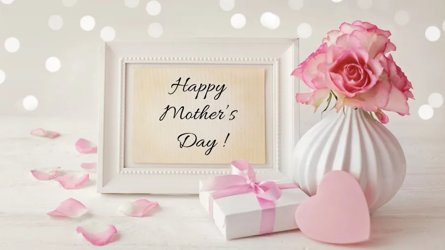 A Mother's Day greeting setup with pink roses, a gift, a heart, and a framed message on a white surface.