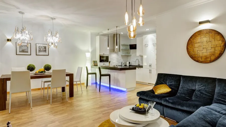 A modern open-concept living and dining area with white walls, a dark velvet sofa, wooden dining table with white chairs, and kitchen with white cabinets and pendant lights.