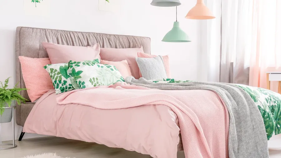 A stylish bedroom interior with cozy pastel pink bedding, throw pillows, throw blanket, lamps hanging from the ceiling, and plants.