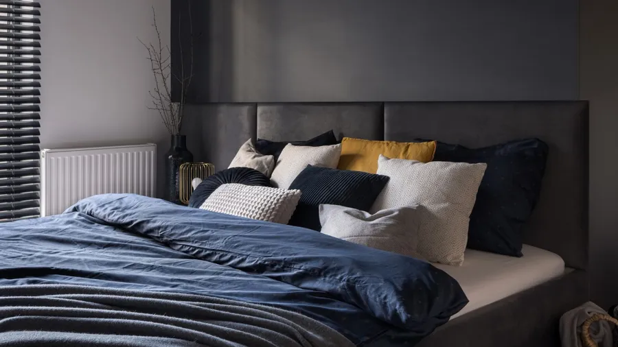 A dark-themed bedroom featuring a plush headboard, various pillows, and window blinds.