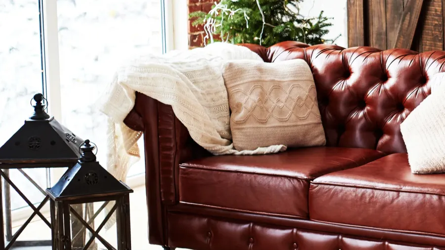 An upholstered leather sofa with textured throw pillows and throw blanket on it near a window.