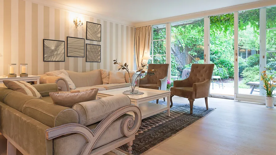 A cozy living room features a beige sofa, decorative pillows, and large glass doors leading to a green garden.