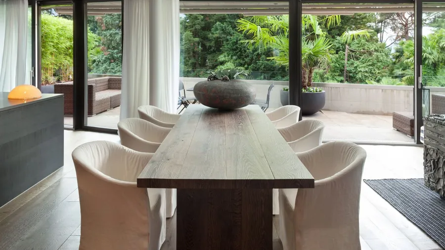 A modern dining area showcases a long wooden table with comfortable chairs and large windows overlooking greenery.
