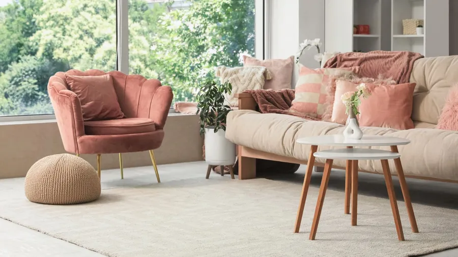 A cozy living room with a pink chair, a beige sofa adorned with pastel pink and beige throw pillows, and a small coffee table with a flower vase.