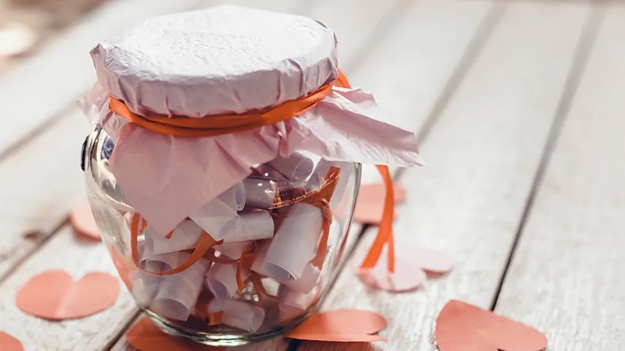 A glass jar filled with crumpled paper and pink paper hearts scattered around on a wooden surface.