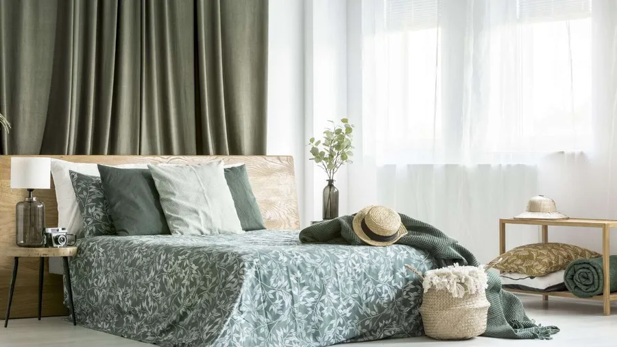 A bedroom interior with a bed with cotton bedding and pillows, wooden table, lamp, flower vase, and woven basket.