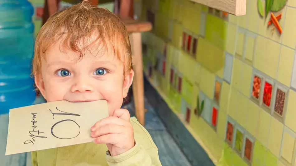 Funny baby in green clothes holds a sign of 10 months old milestone card in his mouth