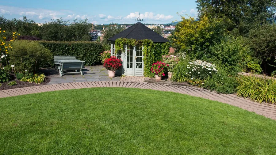 A well-maintained garden with a green lawn, small paved area, gazebo, and colorful flowering plants.