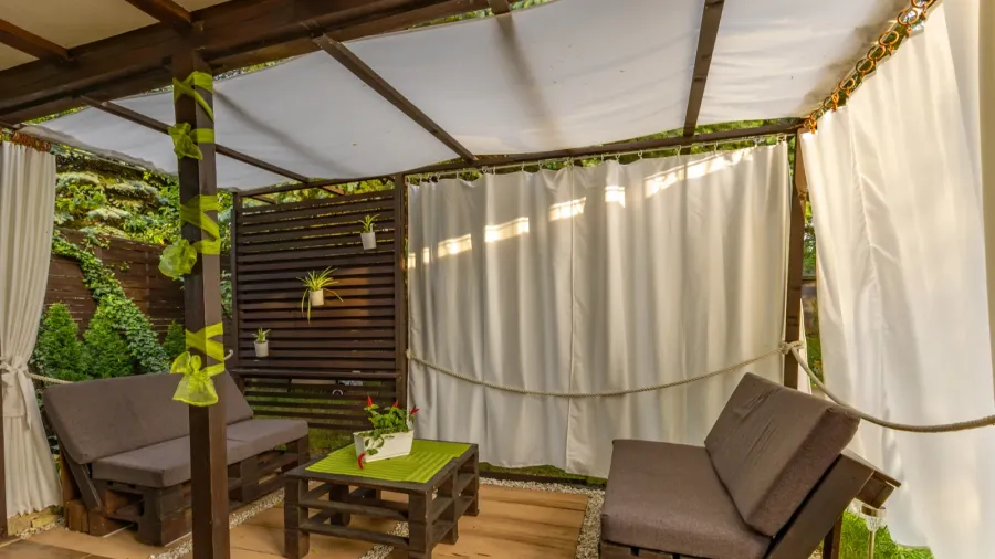 A garden resting area with pallet wooden furniture under a pergola with curtains around it.