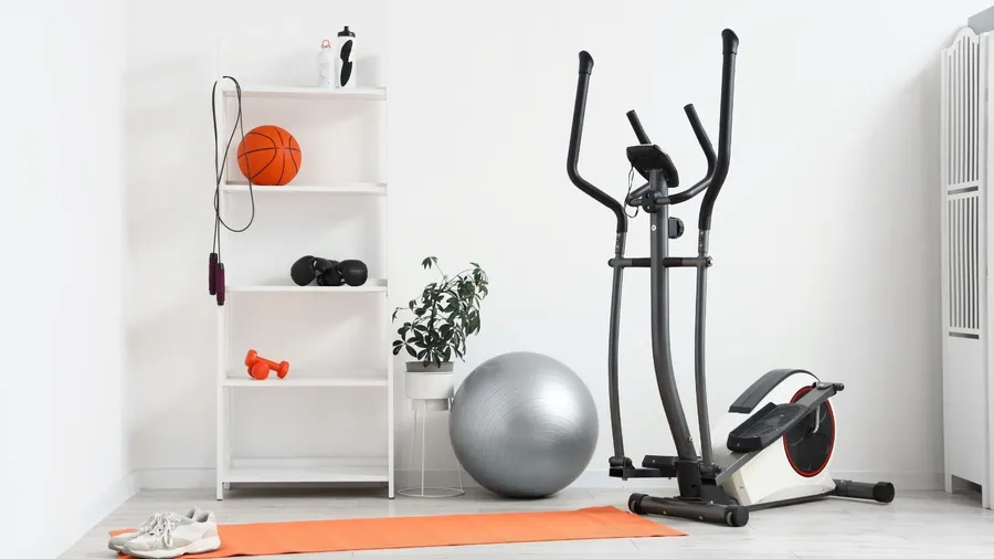 A workout corner in a room with a stability ball, elliptical trainer, mat, and a shelf with sports equipment.