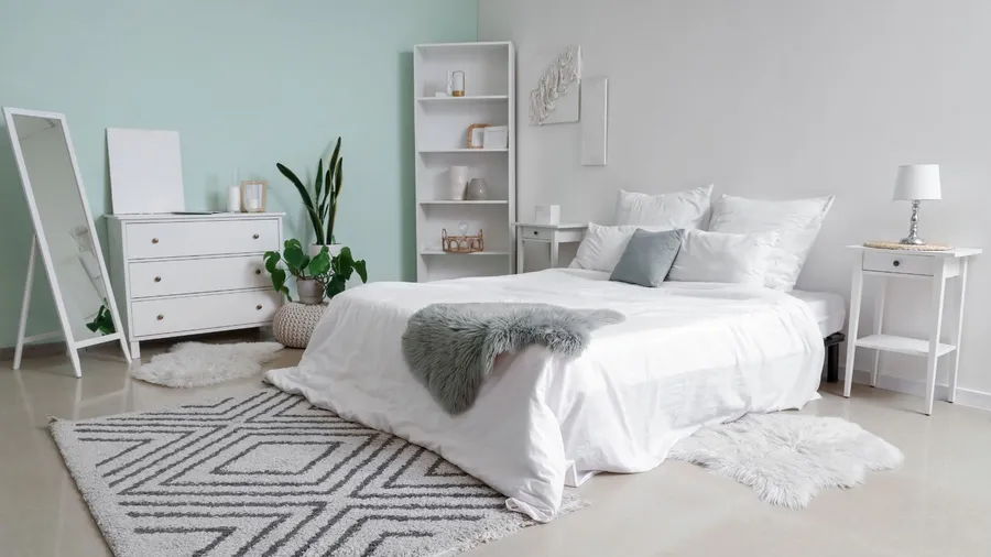 A modern bedroom interior with white bedding, gray and white cushions, gray fur throw, and high-pile shag and faux fur rug on the floor.