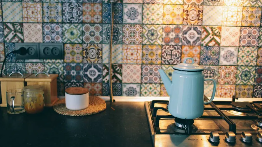 A kitchen interior with kettle on stovetop in front of vibrant mosaic tile backsplash.