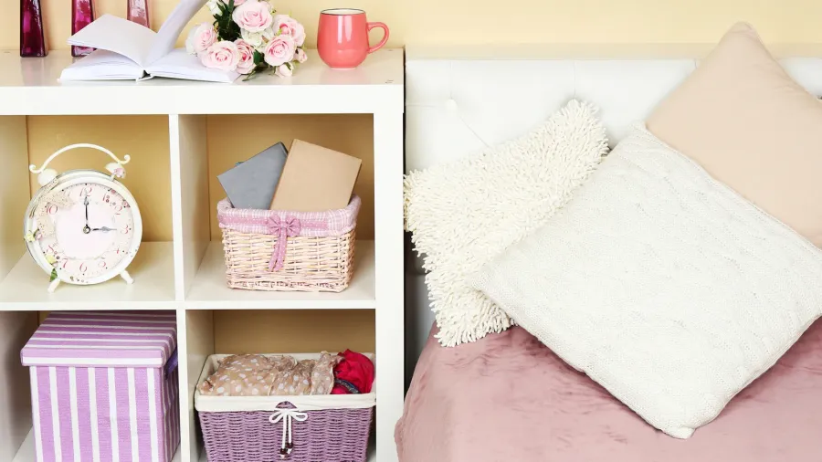 A bedroom interior with a cozy bed with pastel bedding and throw pillows, and a nightstand with shelves containing storage baskets, clock, and book.