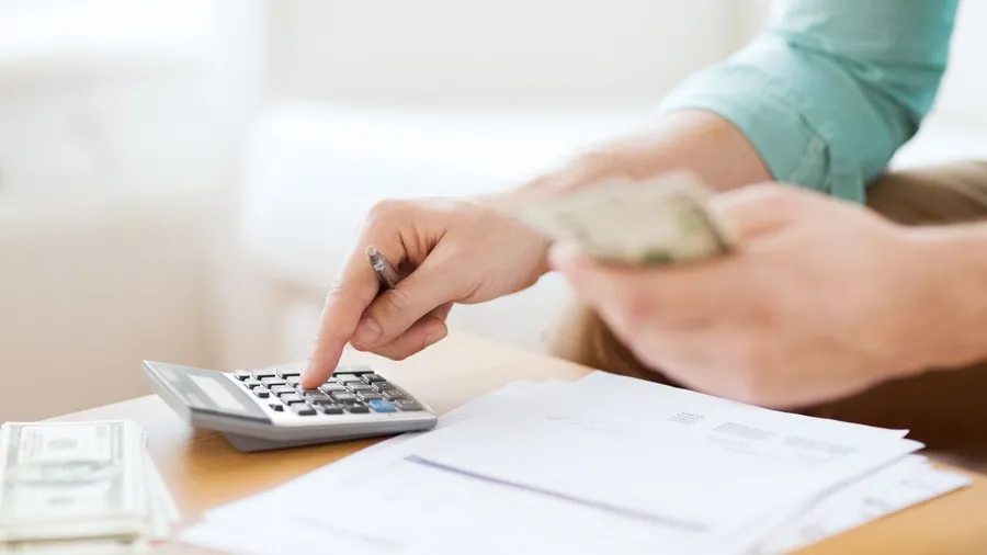 A person using a calculator and making notes on a desk with papers and money.