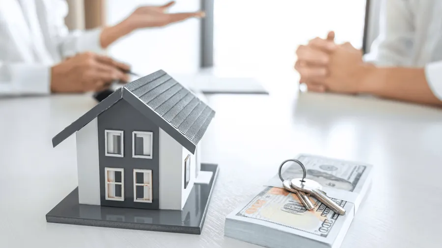 A model house sits on a table next to a stack of cash and keys, with blurred figures of a real estate agent and client in the background.