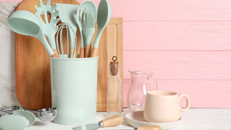 Set of different kitchen utensils on white table against pink wooden background.