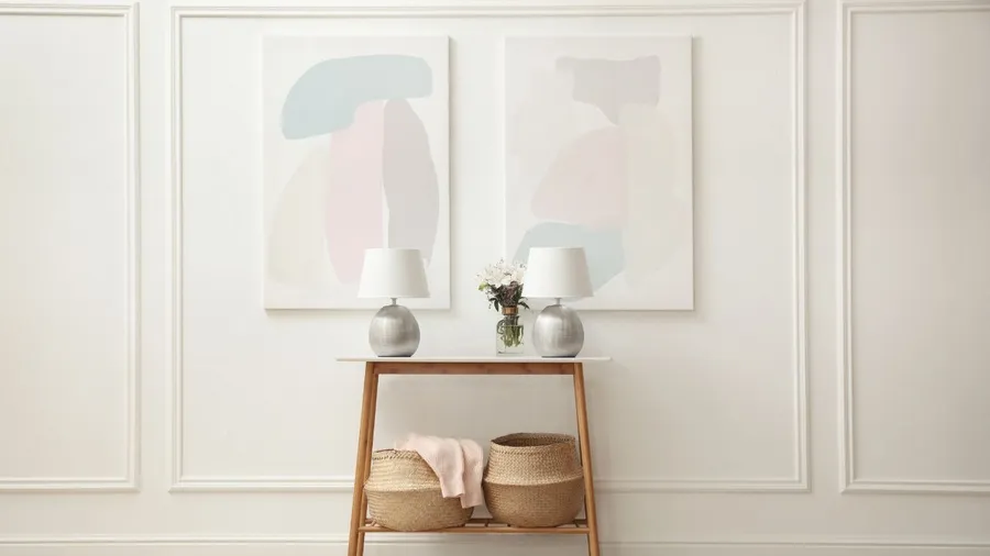 Minimalist hallway or entryway with abstract artwork, white walls, and a wooden console table decorated with lamps and baskets.