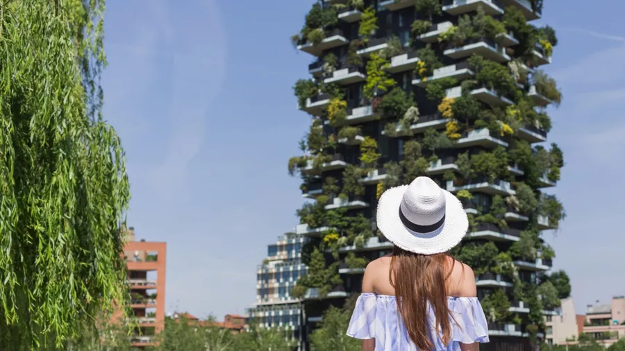 Girl tourist with a sun hat looking at a skyscraper with plant and tree-covered balconies and terraces. Architecture, travel, and lifestyle concepts.