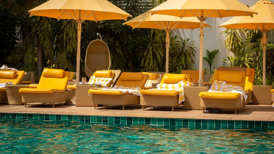 A luxurious swimming pool area in a high end resort or hotel. The pool is surrounded by comfortable yellow lounge chairs, green tropical plants, and modern cane furniture for a spacious elegant look