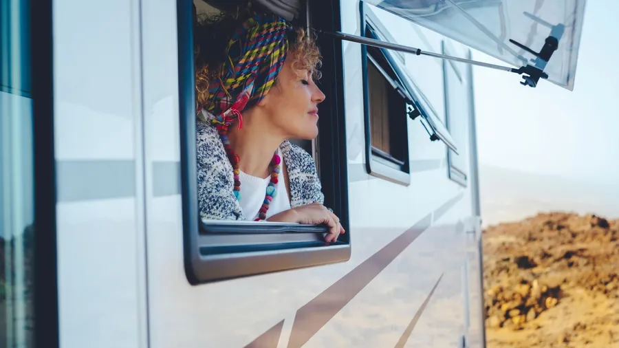 Camper in a desert setting with a curly-haired woman with a scarf on her head leaning against the window of the camper. van with tourist stop on the mountain side to get a better view of the view.