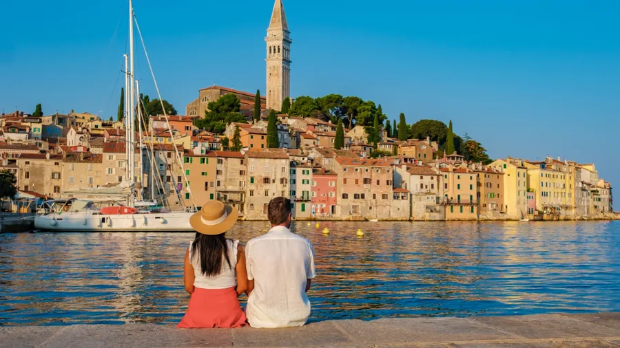 As the sun begins to set, a couple sits by the calm waters in Rovinj, Croatia, admiring the vibrant buildings and reflecting on the day's adventures. A sailboat adds to the picturesque backdrop.