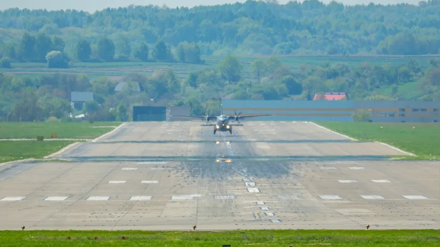 Military aircraft approaches runway during a clear day in a rural airfield surrounded by lush greenery and distant hills