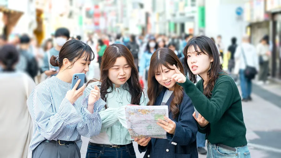 Four young women stand on bustling street, discussing directions with paper map and smartphone in hand.