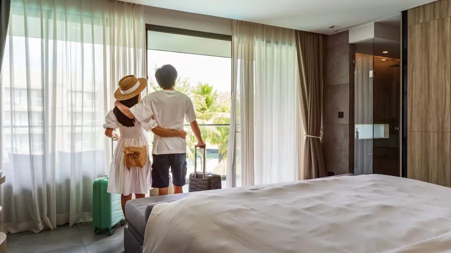 Young couple traveler opening the curtains and looking at the view from the window of a hotel room while on summer vacation, Travel lifestyle concept