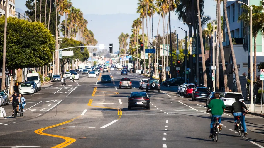 2024-11-15 Los Angeles USA A wide city street in Los Angeles lined with palm trees, featuring cars, cyclists, and clear blue skies, depicting a sunny urban environment.