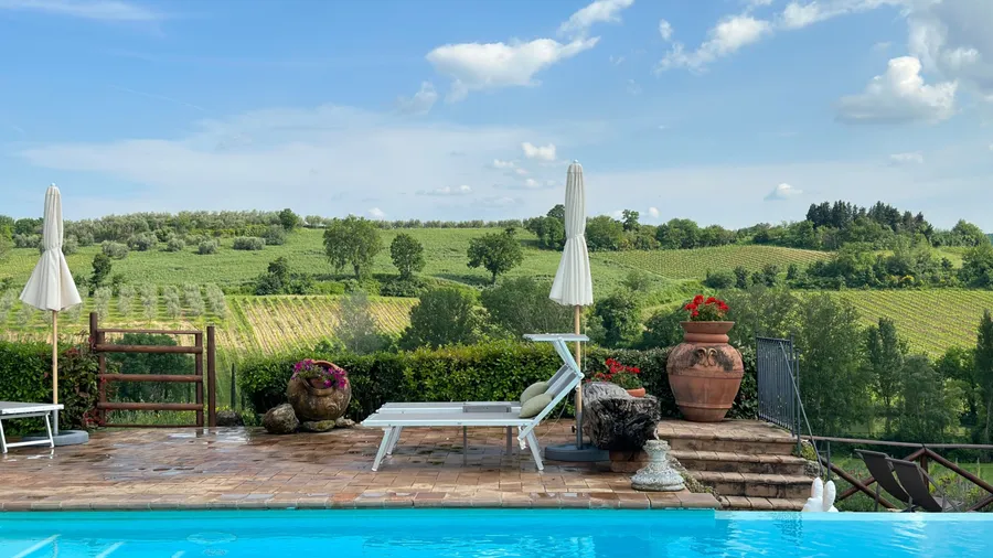 Infinity pool overlooking Tuscan vineyard landscape with rolling green hills and blue summer sky. Terracotta planters and white loungers create perfect Italian villa atmosphere by poolside.