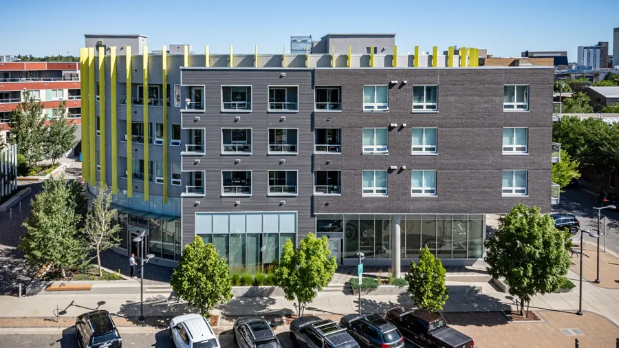 Large building with a yellow trim sits in front of a parking lot. The parking lot is full of cars, including a black car and a white car. The building has a modern design and he is a hotel
