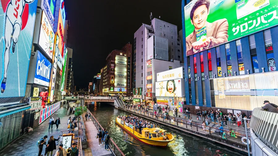 Dotonbori, Osaka, Japan. March 23, 2023. View from the Ebisu bridge at night of a tourist filled cruise boat on the river with the famous Glico Running Man neon display at Dotonbori, Osaka.