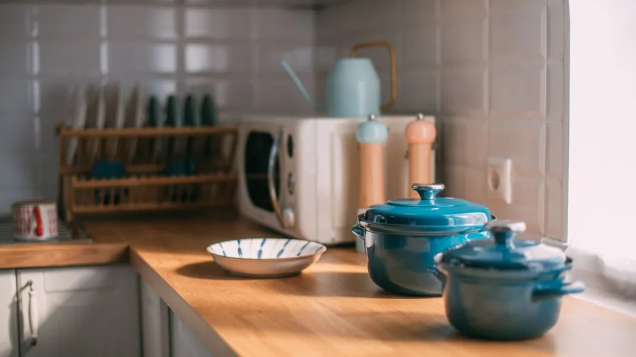 A kitchen interior with wooden countertop with microwave oven, dish rack, and colorful Dutch ovens.