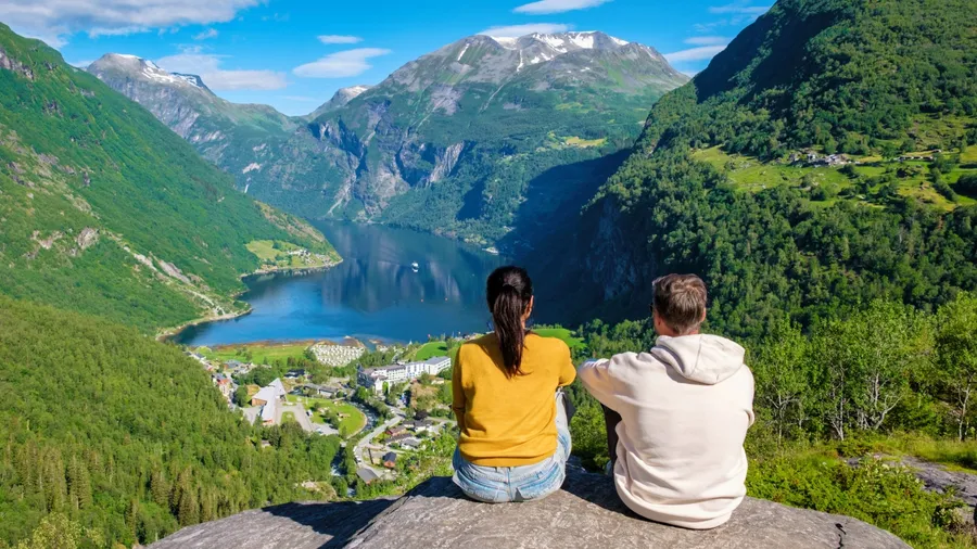 Two people sit on a rocky ledge overlooking a stunning Norwegian fjord, surrounded by lush green mountains and a clear blue sky. Geiranger Fjord Norway. A diverse couple of men and women on vacation