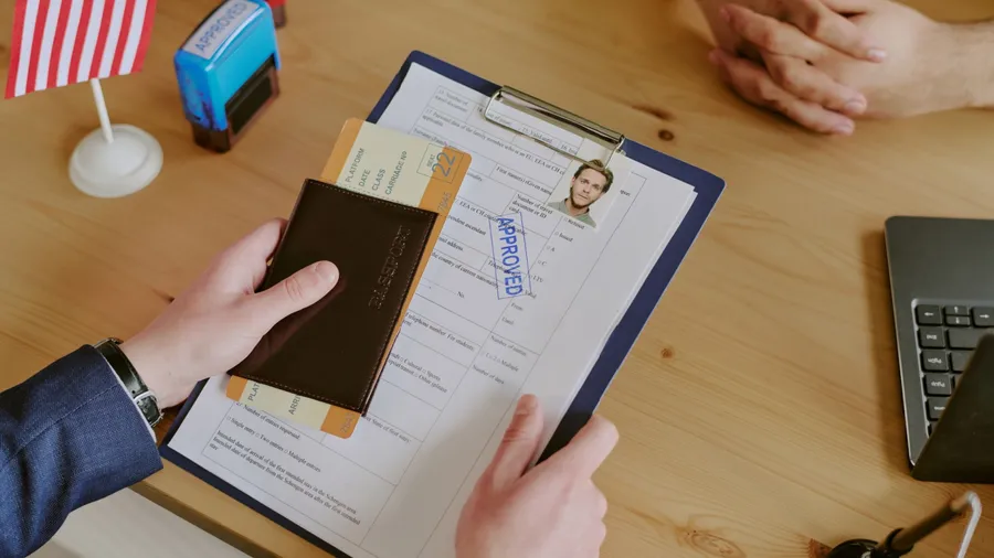 Handing passport and documents across table during visa interview. American flag on table alongside laptop, envelope, and clipboard with approved form visible