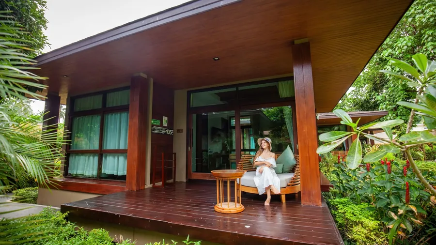 woman in white dress and hat sits near a traditional wooden villa. The tropical paradise of Thailand provides a stunning backdrop for a relaxing vacation. Concept of travel and summer vacation.