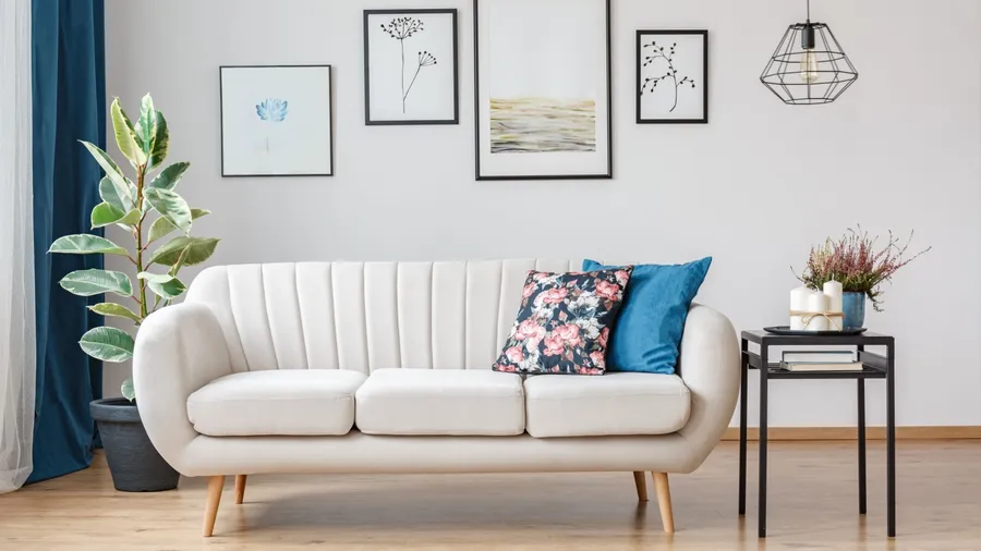A living room interior with plants, side table, ceiling lamp, frames on the wall, and a white sofa with wooden legs.