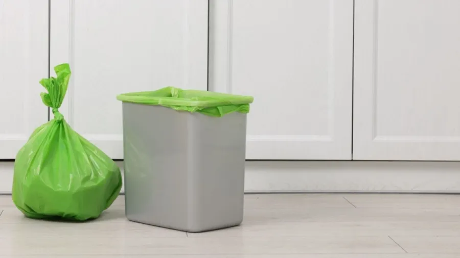 A gray trash can with a green liner beside a bag of green garbage.