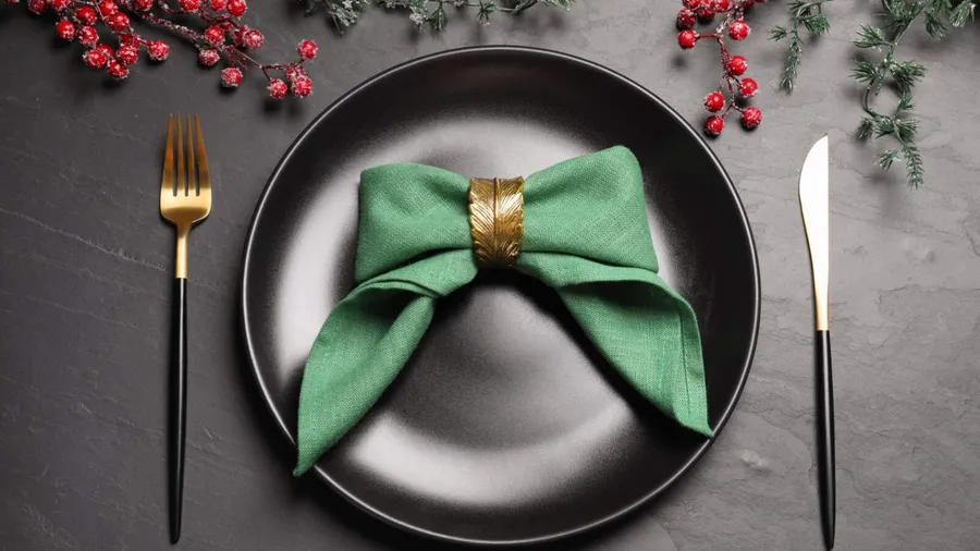 A green napkin with a golden napkin ring on a black plate with utensils on the side on a black dining table.