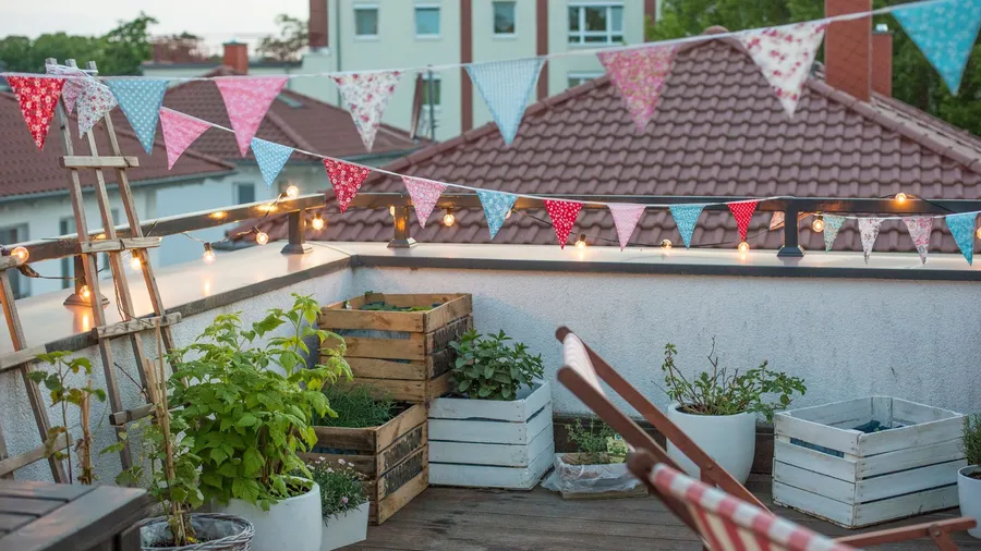 Colorful fabric bunting and string lights decorate a cozy rooftop garden with wooden planters and lounge seating.